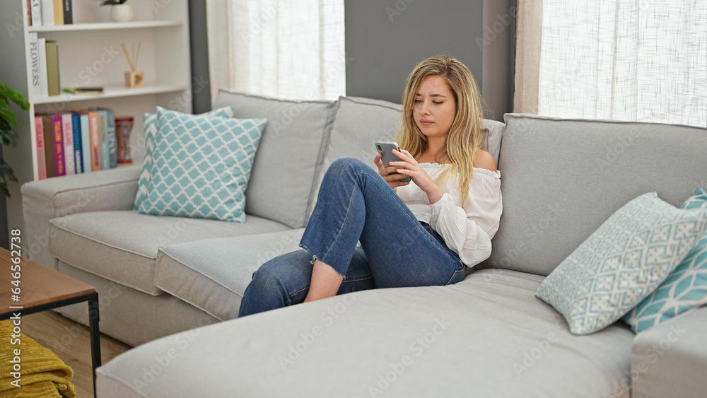 Young blonde woman using smartphone sitting on sofa thinking at home