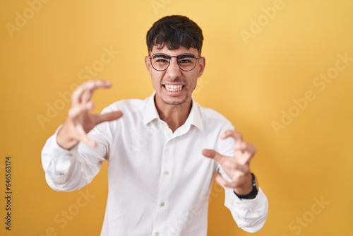 Young hispanic man standing over yellow background shouting frustrated with rage, hands trying to strangle, yelling mad © Krakenimages.com