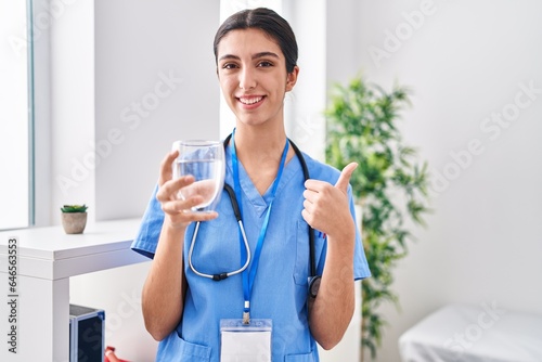 Young beautiful woman wearing doctor uniform and stethoscope drinking water smiling happy and positive, thumb up doing excellent and approval sign