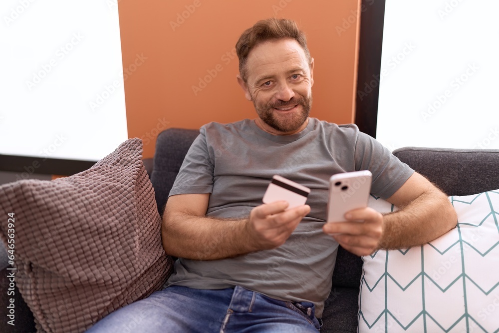 Middle age man using smartphone and credit card sitting on sofa at home Middle age man using smartphone and credit card sitting on sofa at home