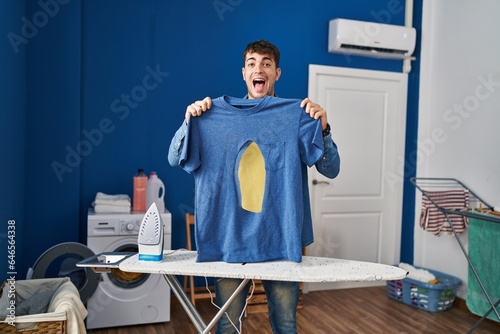 Young hispanic man ironing holding burned iron shirt at laundry room celebrating crazy and amazed for success with open eyes screaming excited.