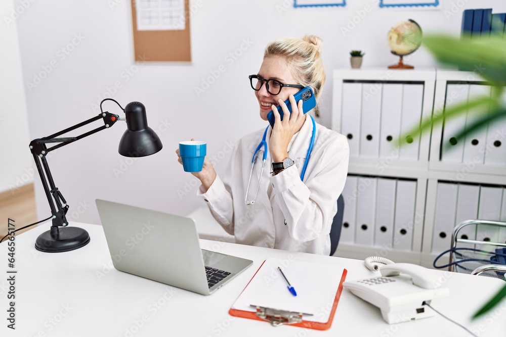 Young blonde woman doctor talking on smartphone drinking coffee at clinic Young blonde woman doctor talking on smartphone drinking coffee at clinic