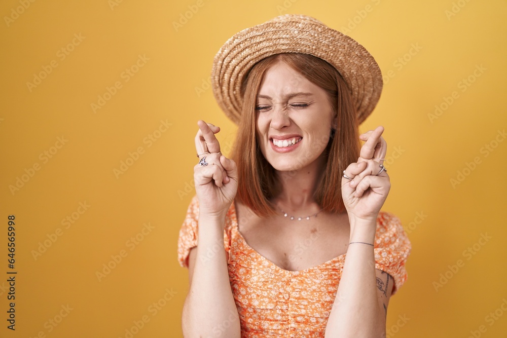 Young redhead woman standing over yellow background wearing summer hat gesturing finger crossed smiling with hope and eyes closed. luck and superstitious concept.