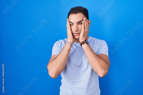 Young hispanic man standing over blue background tired hands covering face, depression and sadness, upset and irritated for problem