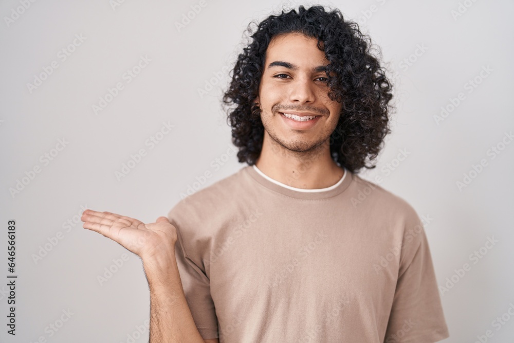 Hispanic man with curly hair standing over white background smiling cheerful presenting and pointing with palm of hand looking at the camera. Hispanic man with curly hair standing over white background smiling cheerful presenting and pointing with palm of hand looking at the camera.