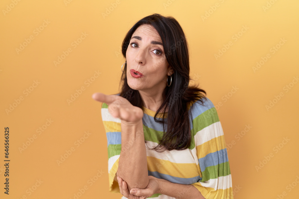 Middle age hispanic woman standing over yellow background looking at the camera blowing a kiss with hand on air being lovely and sexy. love expression. Middle age hispanic woman standing over yellow background looking at the camera blowing a kiss with hand on air being lovely and sexy. love expression.