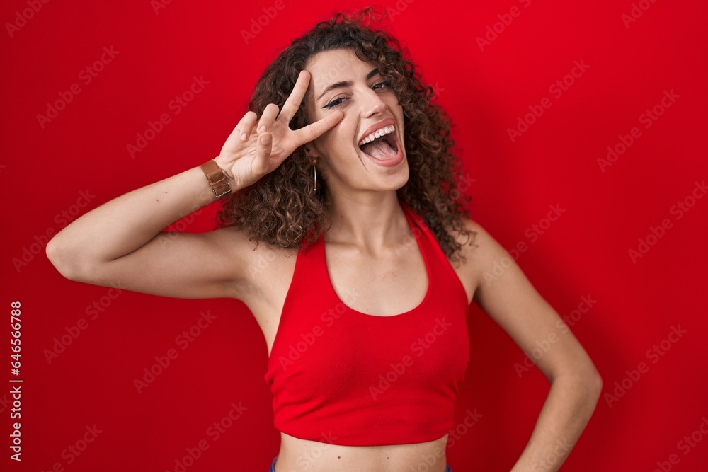 Hispanic woman with curly hair standing over red background doing peace symbol with fingers over face, smiling cheerful showing victory Hispanic woman with curly hair standing over red background doing peace symbol with fingers over face, smiling cheerful showing victory
