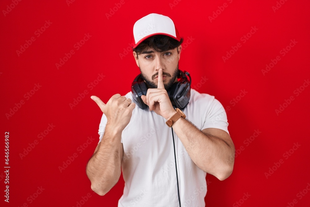 Hispanic man with beard wearing gamer hat and headphones asking to be quiet with finger on lips pointing with hand to the side. silence and secret concept. Hispanic man with beard wearing gamer hat and headphones asking to be quiet with finger on lips pointing with hand to the side. silence and secret concept.