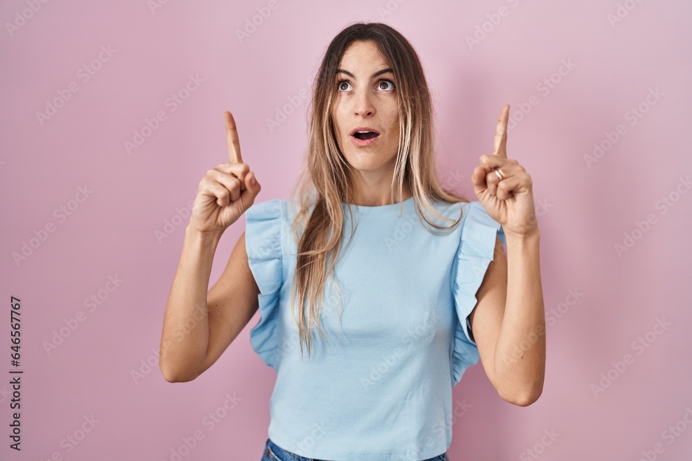 Young hispanic woman standing over pink background amazed and surprised looking up and pointing with fingers and raised arms. Young hispanic woman standing over pink background amazed and surprised looking up and pointing with fingers and raised arms.