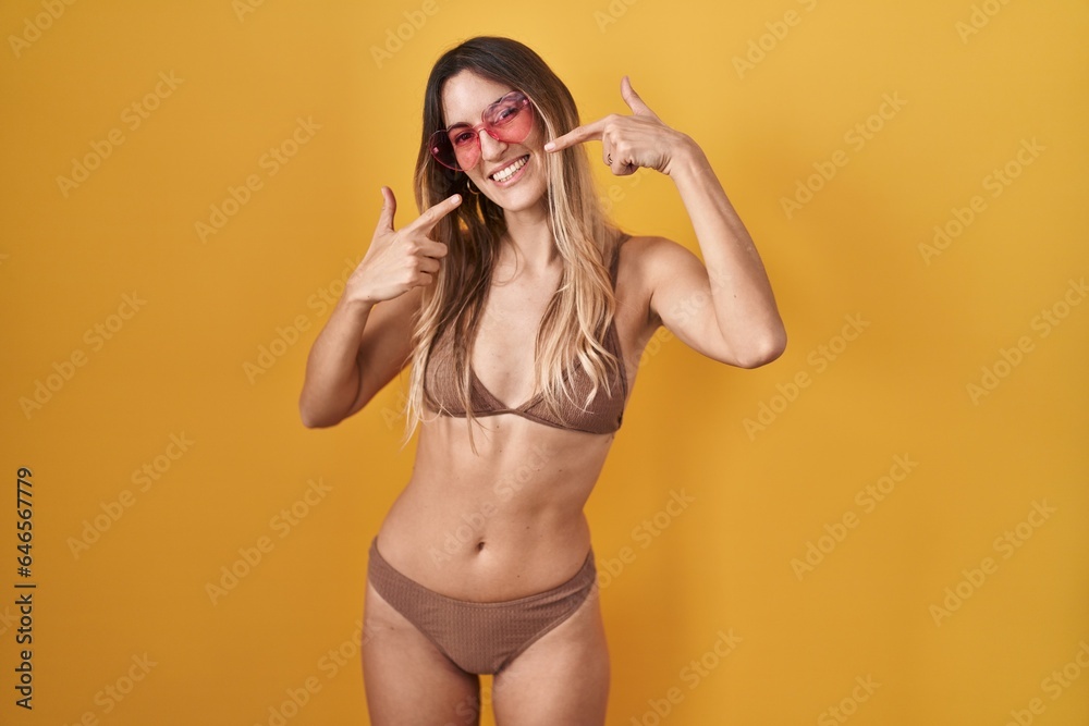 Young hispanic woman wearing bikini over yellow background smiling cheerful showing and pointing with fingers teeth and mouth. dental health concept. Young hispanic woman wearing bikini over yellow background smiling cheerful showing and pointing with fingers teeth and mouth. dental health concept.