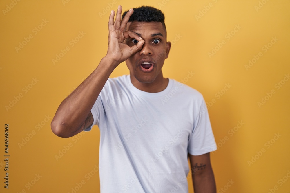Young hispanic man standing over yellow background doing ok gesture shocked with surprised face, eye looking through fingers. unbelieving expression. Young hispanic man standing over yellow background doing ok gesture shocked with surprised face, eye looking through fingers. unbelieving expression.