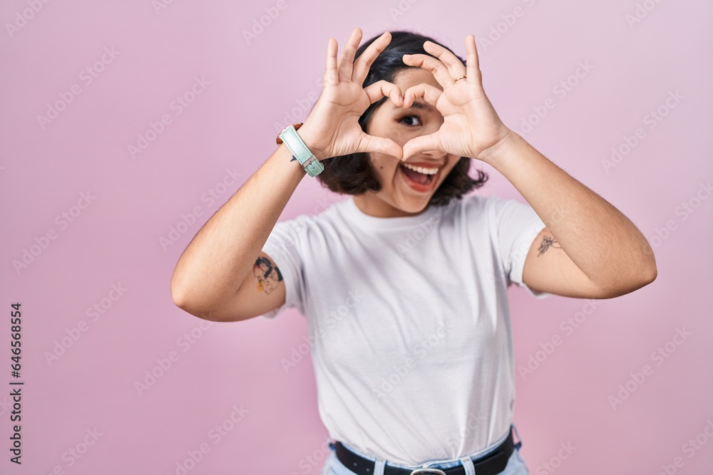 Young hispanic woman wearing casual white t shirt over pink background doing heart shape with hand and fingers smiling looking through sign Young hispanic woman wearing casual white t shirt over pink background doing heart shape with hand and fingers smiling looking through sign