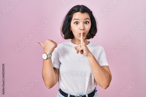 Young hispanic woman wearing casual white t shirt over pink background asking to be quiet with finger on lips pointing with hand to the side. silence and secret concept.