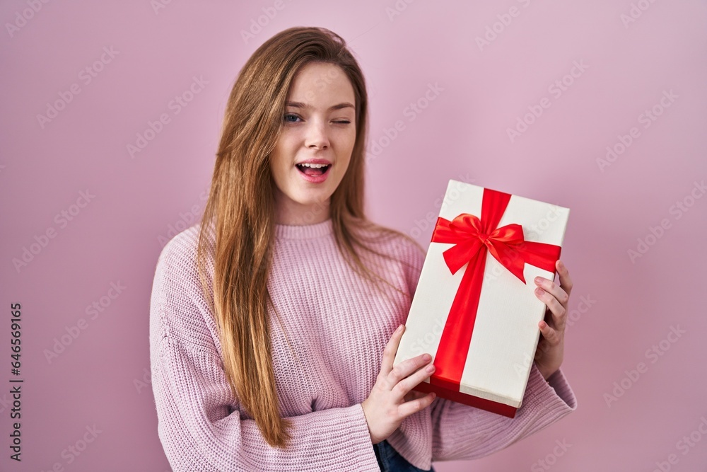Young caucasian woman holding gift winking looking at the camera with sexy expression, cheerful and happy face. Young caucasian woman holding gift winking looking at the camera with sexy expression, cheerful and happy face.