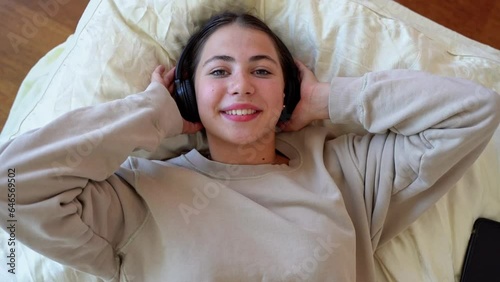 Young teen girl woman with headphones in lying bedroom smiling listen to music radio podcast, dance, enjoy life. Technology in daily life, Youth culture Top side view photo