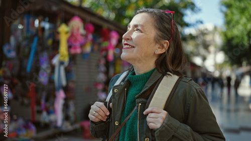 Mature hispanic woman with grey hair tourist wearing backpack smiling looking to the side at street market photo