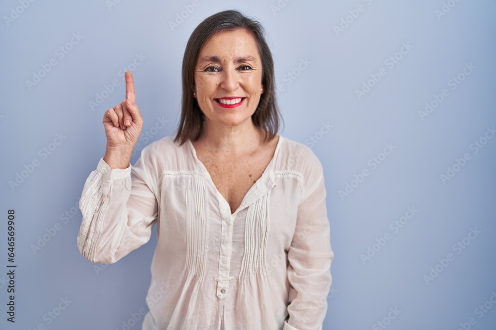 Middle age hispanic woman standing over blue background showing and pointing up with finger number one while smiling confident and happy. Middle age hispanic woman standing over blue background showing and pointing up with finger number one while smiling confident and happy.
