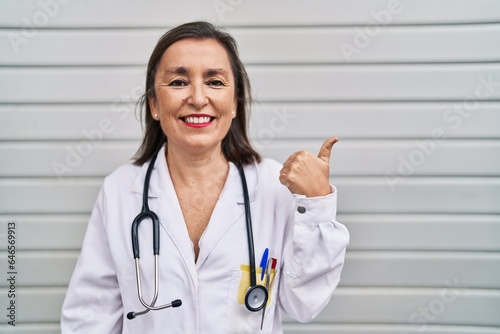 Middle age hispanic woman wearing doctor uniform and stethoscope pointing thumb up to the side smiling happy with open mouth