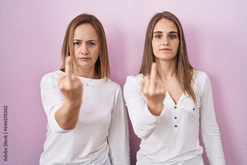 Middle age mother and young daughter standing over pink background showing middle finger, impolite and rude fuck off expression Middle age mother and young daughter standing over pink background showing middle finger, impolite and rude fuck off expression