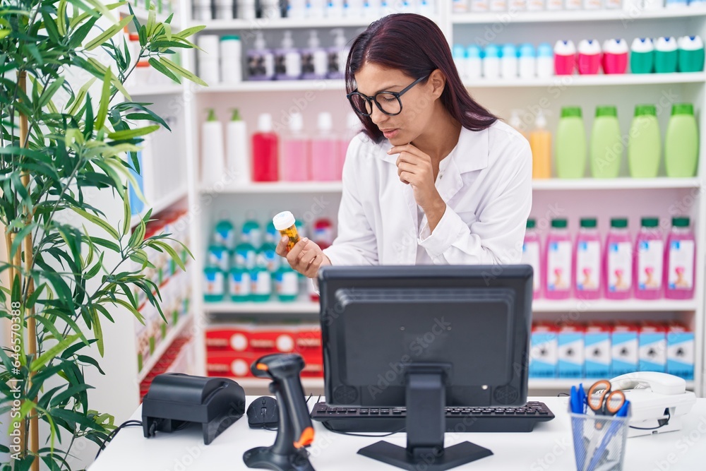 Young beautiful hispanic woman pharmacist using computer holding pills bottle at pharmacy Young beautiful hispanic woman pharmacist using computer holding pills bottle at pharmacy