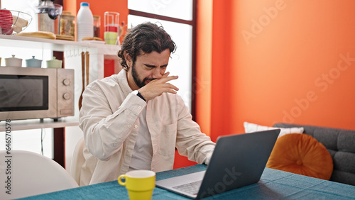 Young hispanic man using laptop sneezing at dinning room