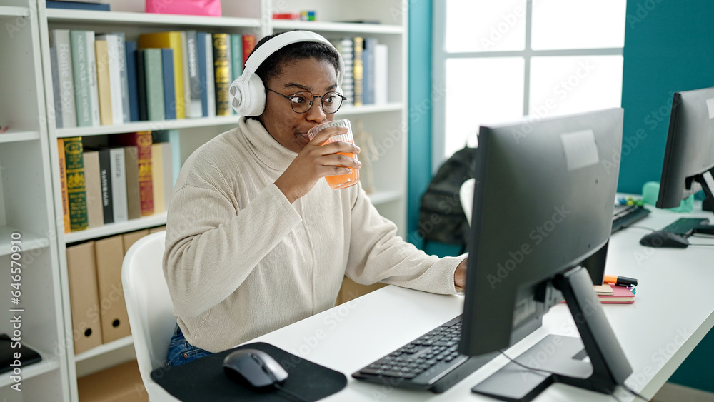African american woman student using computer and headphones drinking orange juice at library university African american woman student using computer and headphones drinking orange juice at library university