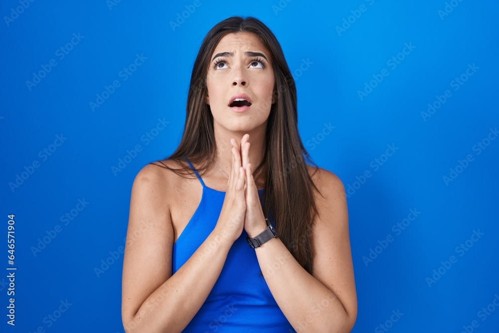 Hispanic woman standing over blue background begging and praying with hands together with hope expression on face very emotional and worried. begging. Hispanic woman standing over blue background begging and praying with hands together with hope expression on face very emotional and worried. begging.
