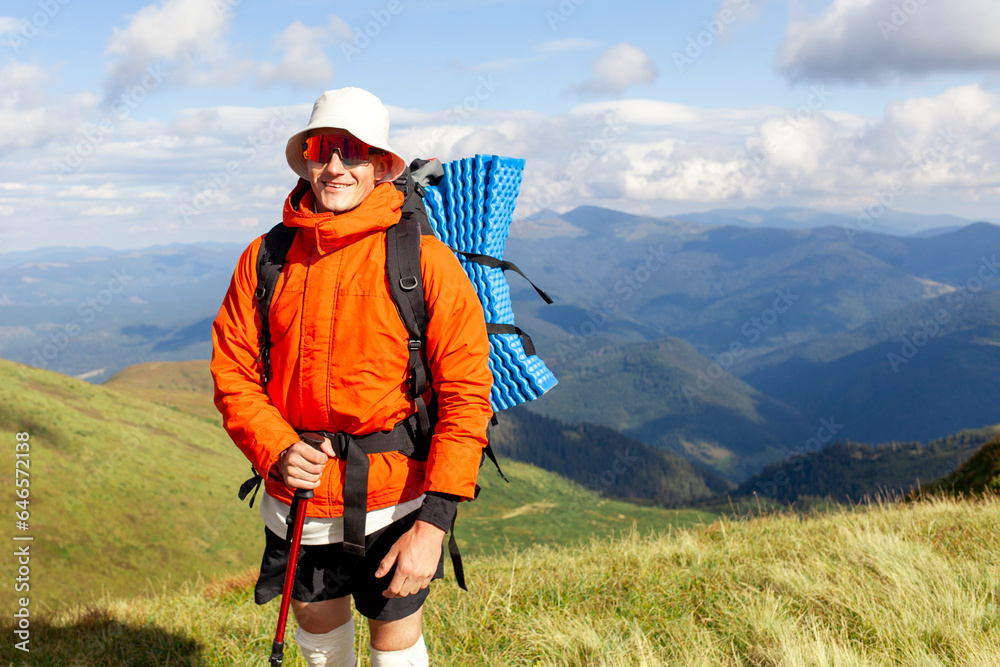 young man in orange jacket and glasses stands on the top of mountain with backpack and trekking stick
