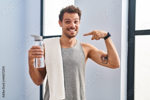 Young hispanic man wearing sportswear drinking water looking confident with smile on face, pointing oneself with fingers proud and happy.