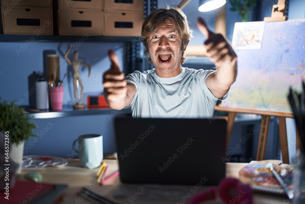 Middle age man sitting at art studio with laptop at night approving doing positive gesture with hand, thumbs up smiling and happy for success. winner gesture. Middle age man sitting at art studio with laptop at night approving doing positive gesture with hand, thumbs up smiling and happy for success. winner gesture.