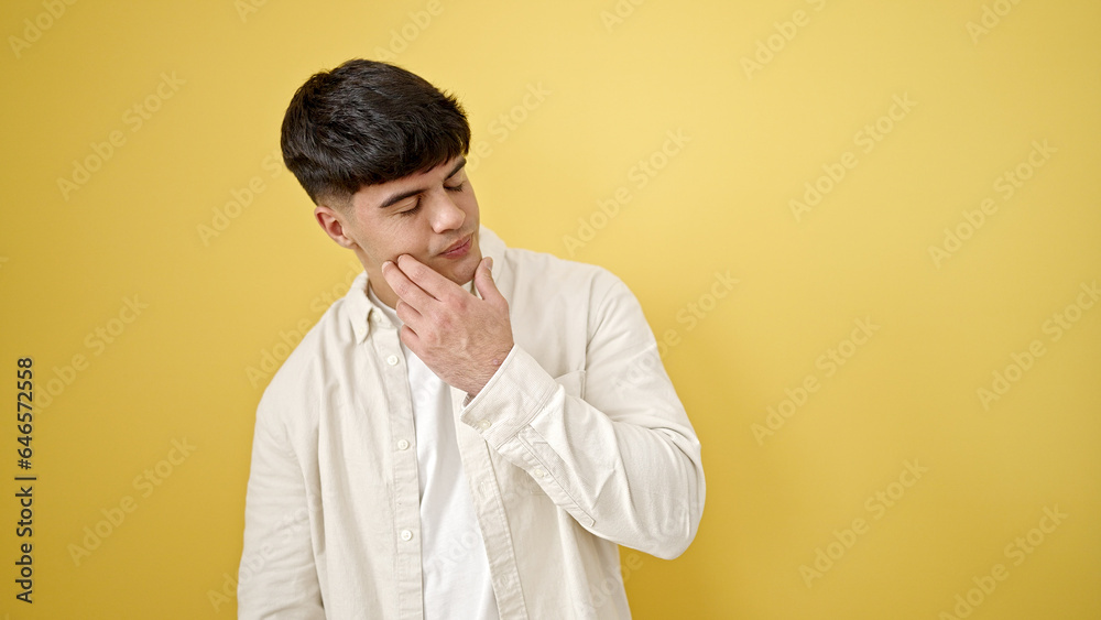 Young hispanic man standing with serious expression touching face over isolated yellow background