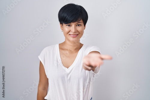 Young asian woman with short hair standing over isolated background smiling cheerful offering palm hand giving assistance and acceptance.