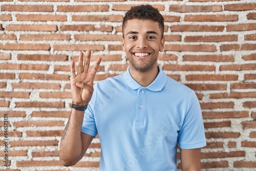 Brazilian young man standing over brick wall showing and pointing up with fingers number four while smiling confident and happy.