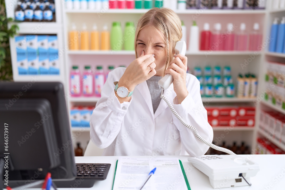 Young caucasian woman working at pharmacy drugstore speaking on the telephone smelling something stinky and disgusting, intolerable smell, holding breath with fingers on nose. bad smell Young caucasian woman working at pharmacy drugstore speaking on the telephone smelling something stinky and disgusting, intolerable smell, holding breath with fingers on nose. bad smell