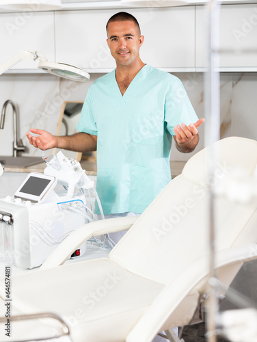 Portrait of positive male doctor cosmetologist wearing overall showing equipment of clinic before procedure