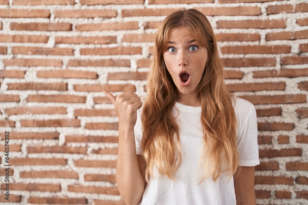 Young caucasian woman standing over bricks wall surprised pointing with hand finger to the side, open mouth amazed expression. Young caucasian woman standing over bricks wall surprised pointing with hand finger to the side, open mouth amazed expression.