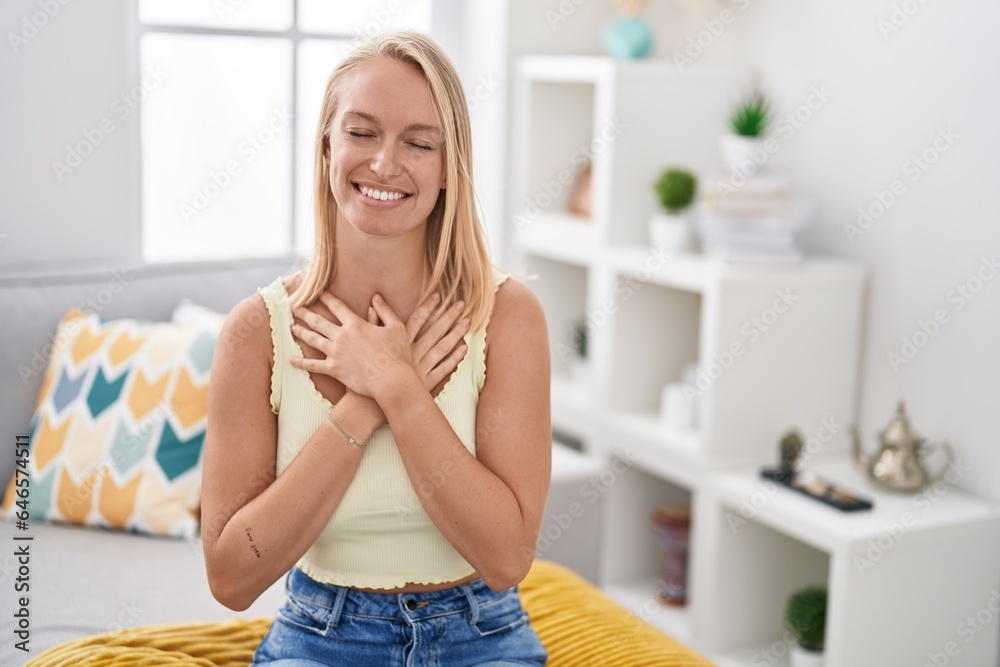 Young blonde woman sitting on sofa with hands on heart at home