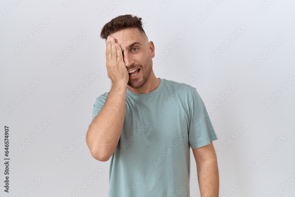 Hispanic man with beard standing over white background covering one eye with hand, confident smile on face and surprise emotion. Hispanic man with beard standing over white background covering one eye with hand, confident smile on face and surprise emotion.