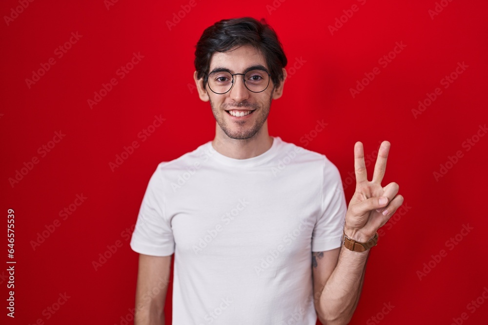 Naklejka premium Young hispanic man standing over red background showing and pointing up with fingers number two while smiling confident and happy.