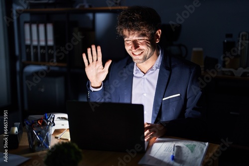 Hispanic young man working at the office at night showing and pointing up with fingers number five while smiling confident and happy.