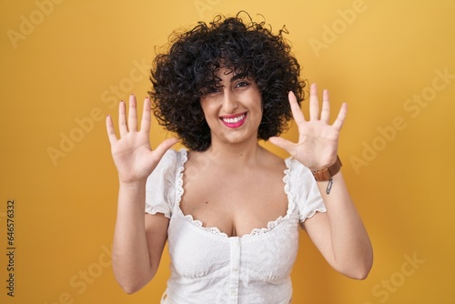 Young brunette woman with curly hair standing over yellow background showing and pointing up with fingers number ten while smiling confident and happy.