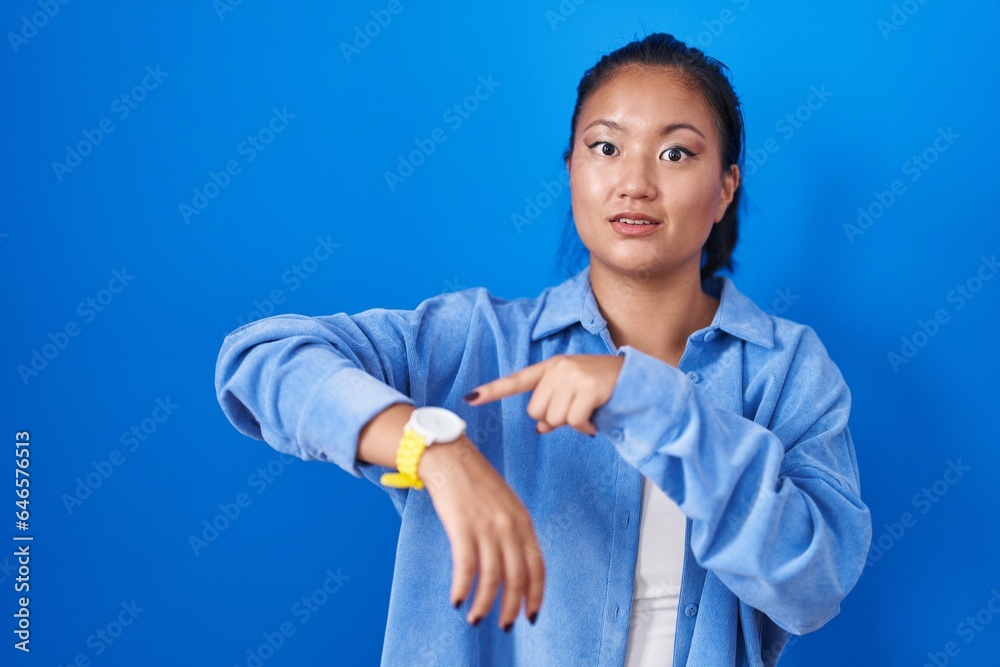 Asian young woman standing over blue background in hurry pointing to watch time, impatience, upset and angry for deadline delay