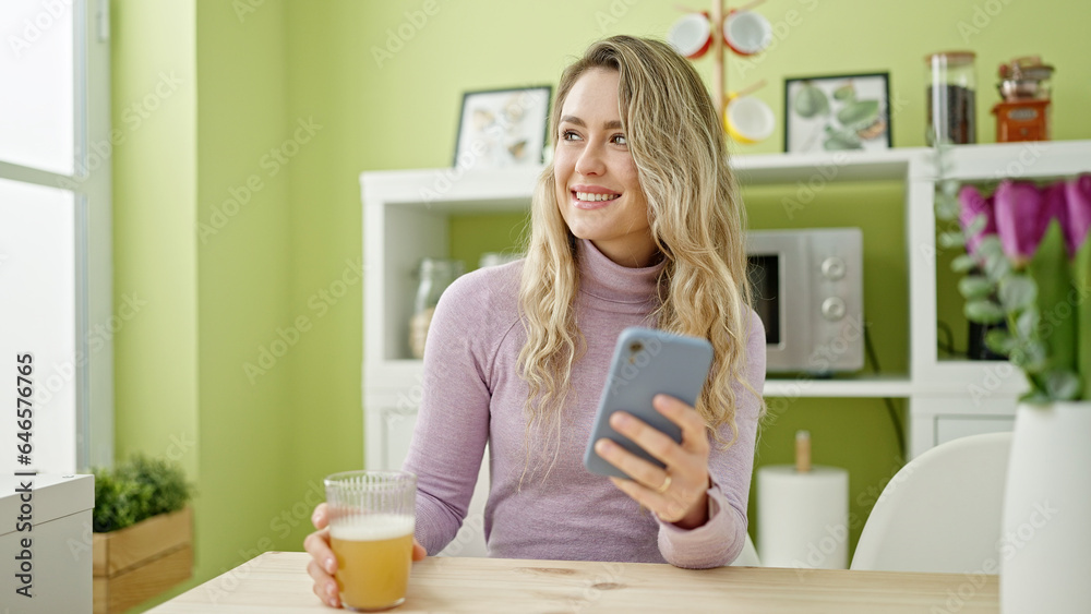 Young blonde woman holding glass of orange juice using smartphone at dinning room Young blonde woman holding glass of orange juice using smartphone at dinning room
