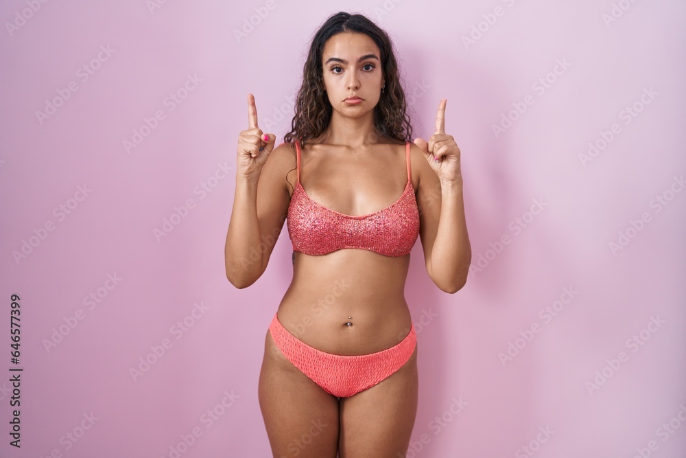 Naklejka premium Young hispanic woman wearing lingerie over pink background pointing up looking sad and upset, indicating direction with fingers, unhappy and depressed.