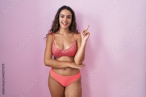 Young hispanic woman wearing lingerie over pink background with a big smile on face, pointing with hand finger to the side looking at the camera.