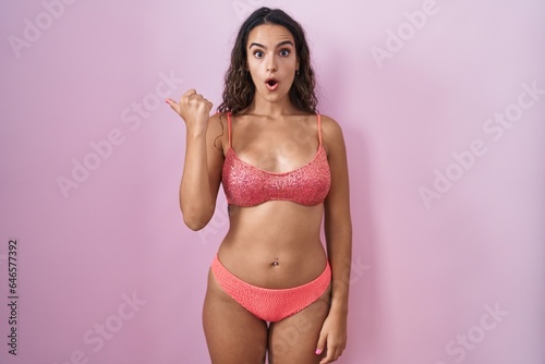 Young hispanic woman wearing lingerie over pink background surprised pointing with hand finger to the side, open mouth amazed expression.
