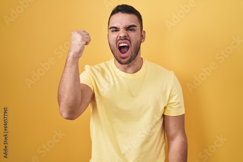 Young hispanic man standing over yellow background angry and mad raising fist frustrated and furious while shouting with anger. rage and aggressive concept. © Krakenimages.com
