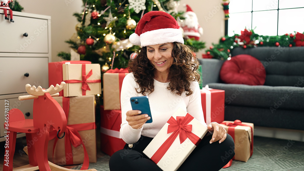 Middle age hispanic woman using smartphone sitting on floor by christmas tree at home Middle age hispanic woman using smartphone sitting on floor by christmas tree at home