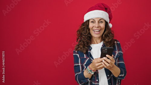 Middle age hispanic woman wearing christmas hat using smartphone over isolated red background