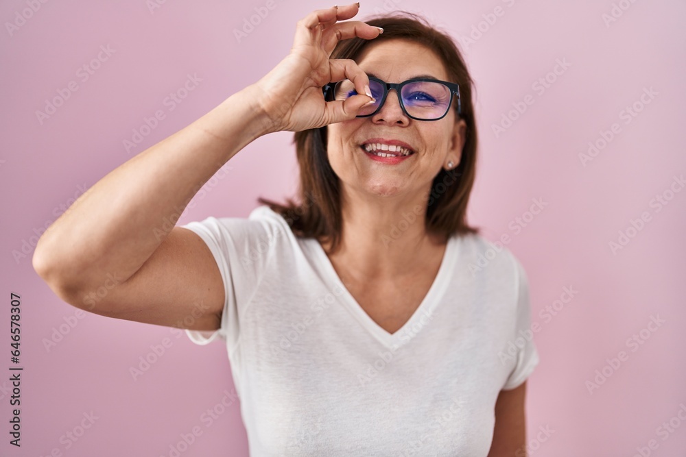 Middle age hispanic woman standing over pink background doing ok gesture with hand smiling, eye looking through fingers with happy face.